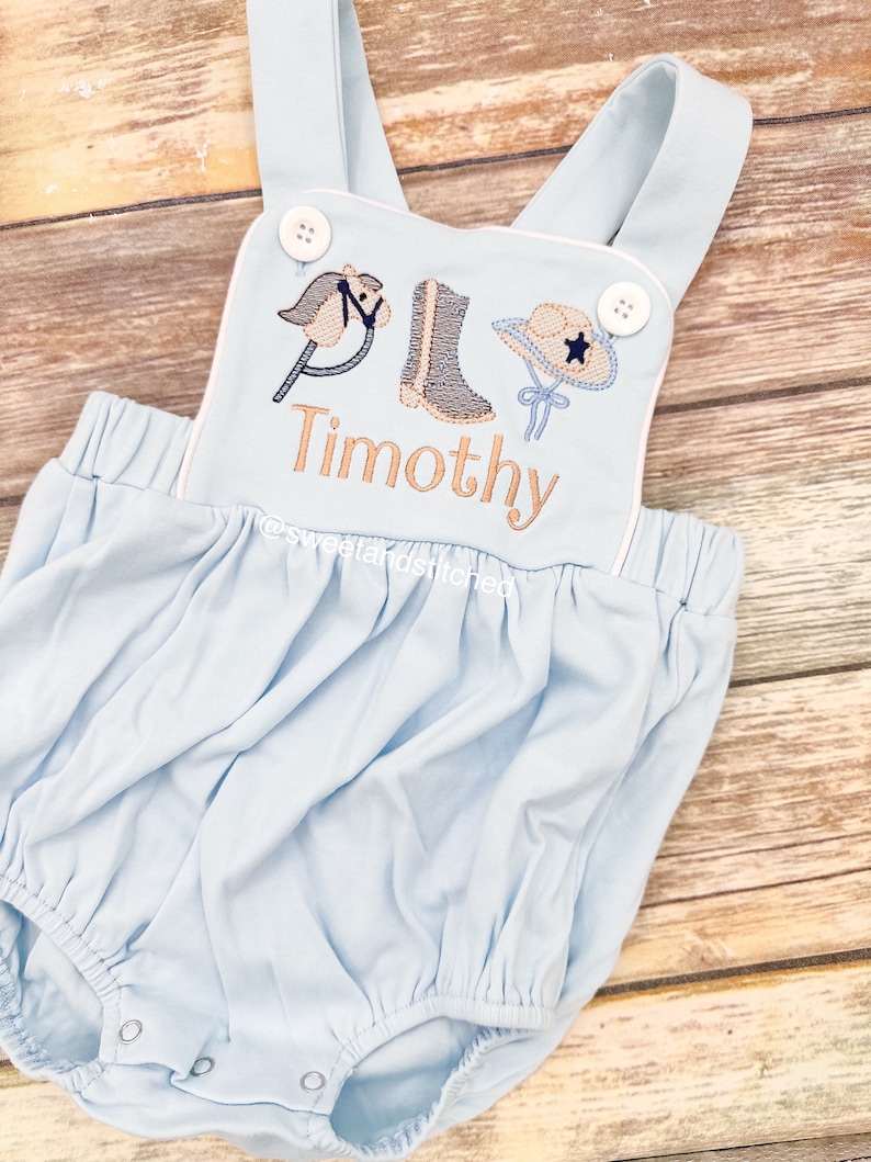 May include: Light blue baby onesie with white straps and white trim. The onesie has an embroidered design of a rocking horse, cowboy boot, and cowboy hat with a star. The name "Timothy" is embroidered below the design.