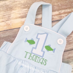May include: Light blue overalls with white trim and a white number one with green fish on either side. The name "Thomas" is embroidered in green below the number one. The text "@sweetandstitched" is embroidered in green below the name.