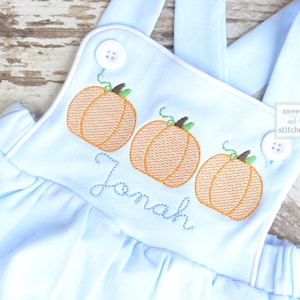 May include: Light blue baby overalls with a white trim and three embroidered pumpkins. The overalls have a button closure and the name "Jonah" is embroidered below the pumpkins.