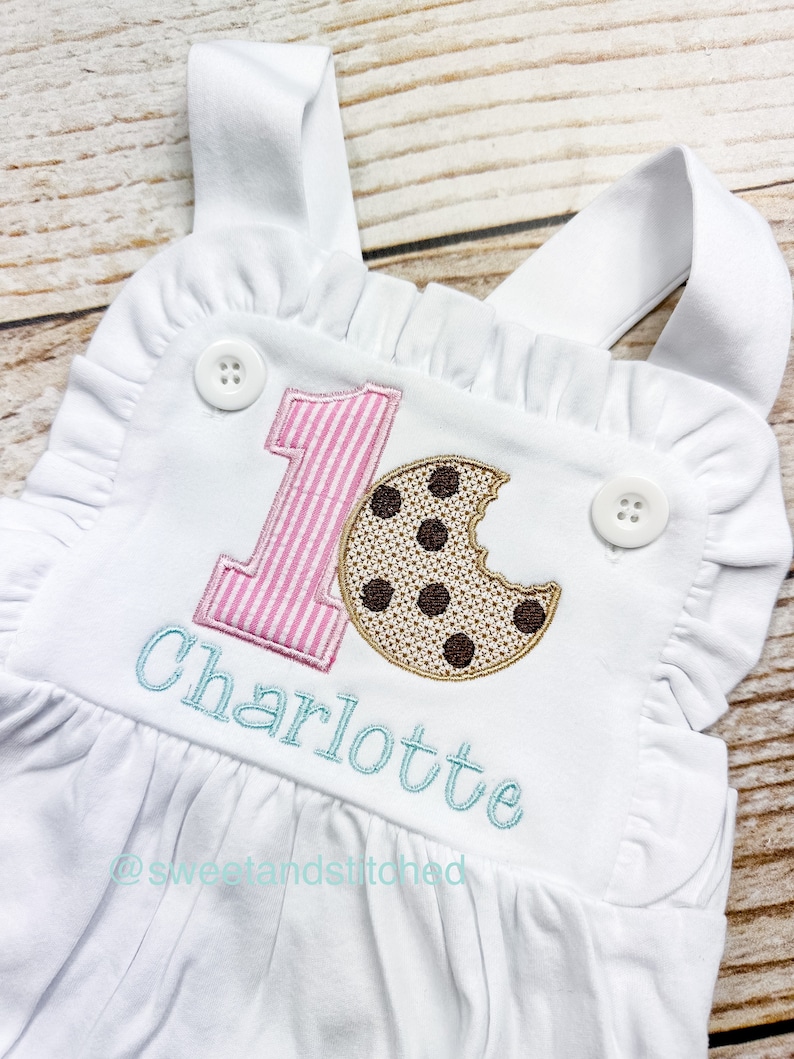 K&ouml;nnte beinhalten: Wei&szlig;er Baby-Strampler mit pink-wei&szlig; gestreiftem Nummer eins, einem braunen Schokoladenkeks und dem Namen Charlotte in Blau gestickt. Der Strampler hat R&uuml;schen an den R&auml;ndern und Tr&auml;ger.