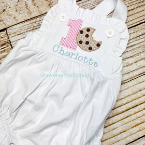 K&ouml;nnte beinhalten: Wei&szlig;er Baby-Strampler mit pink-wei&szlig; gestreifter Nummer eins und einem braunen Schokoladenkeks mit einem Bissen heraus. Der Strampler ist mit dem Namen "Charlotte" und dem Social-Media-Handle "@sweetandstitched" personalisiert.