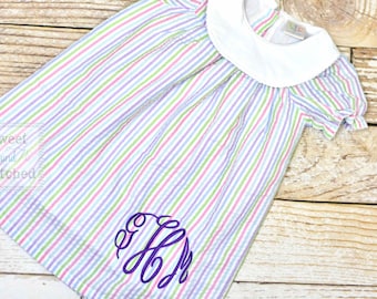 May include: A white and pastel striped dress with a white Peter Pan collar and short sleeves. The dress has a purple monogram on the front.
