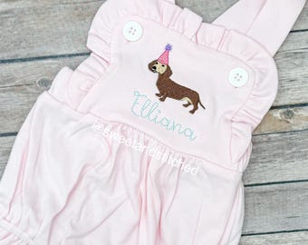 Może przedstawiać: Różowy pajacyk dla dziecka z falbankami i guzikami. Zawiera haftowanego jamnika w czapeczce, imię "Iliana" w skrypcie i tekst "@sweetandstitched".