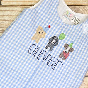 May include: A blue and white gingham baby onesie with a personalized name "Oliver" embroidered on the front. Three embroidered dogs are wearing party hats and one is holding a balloon. The onesie has a small tag that says "sweet & stitched".