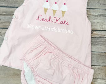 May include: A light pink baby outfit set, featuring a sleeveless top and matching bloomers. The top has an embroidered ice cream cone design and the text "Leah Kate @sweetandstitched". The outfit is made of soft fabric and has button closures.