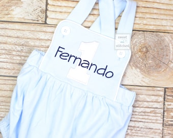May include: Light blue baby overalls with white trim and a white number one with the name "Fernando" embroidered in navy blue.