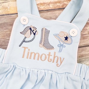 May include: Light blue baby overalls with white piping. The overalls have an embroidered design of a rocking horse, cowboy boot, and cowboy hat. The name "Timothy" is embroidered below the design. The text "@sweetlandstitched" is embroidered below the name.