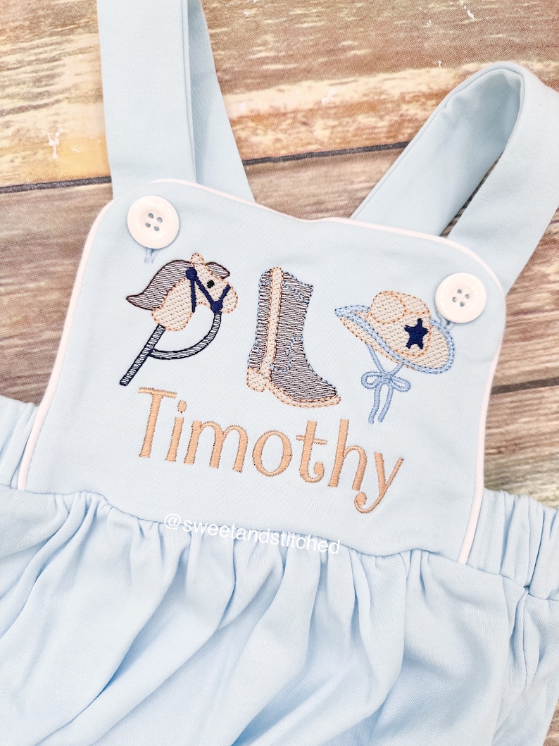 May include: Light blue baby overalls with white piping. The overalls have an embroidered design of a rocking horse, cowboy boot, and cowboy hat. The name "Timothy" is embroidered below the design. The text "@sweetlandstitched" is embroidered below the name.
