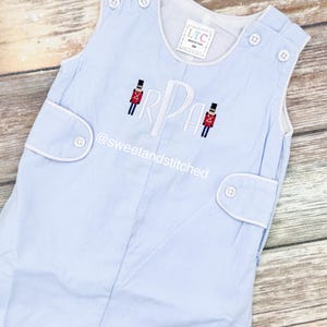 May include: Light blue corduroy romper with white trim and button details. Features embroidered initials and toy soldier figures. The text "@sweetandstitched" is visible on the front. A tag reads "LTC MADE IN USA 9M".