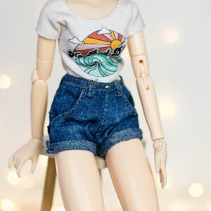May include: A doll with auburn hair styled in two buns, wearing round gold-rimmed glasses, a white t-shirt with a colorful graphic, and denim shorts. The doll is sitting on a small wooden stool.