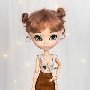 Op de afbeelding: Een modepop met roodbruin haar in twee knotjes. De pop draagt een crèmekleurige top, een bruine corduroy rok en zwarte panty's met patroon. Ze heeft een kleine beige handtas en bruine laarzen. De pop heeft grote groene ogen en staat in een casual pose.