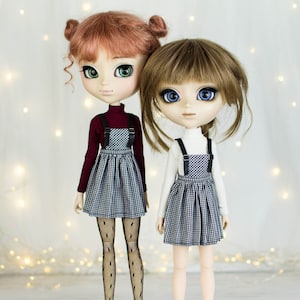 Può includere: Due bambole che indossano abiti a quadri bianchi e neri. La bambola a sinistra ha i capelli rossi e indossa un dolcevita bordeaux. La bambola a destra ha i capelli castani e indossa un dolcevita bianco.