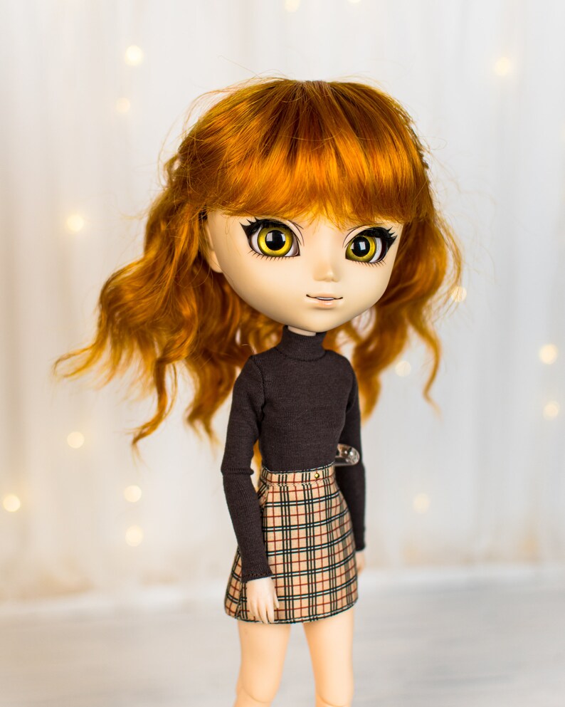 May include: A doll with long, curly, auburn hair, wearing a black turtleneck sweater and a plaid skirt. The doll has large, yellow eyes and a neutral expression.