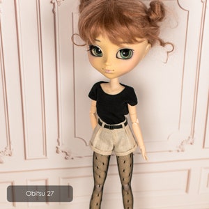May include: A doll with brown hair styled in two buns, wearing a black short-sleeved shirt, beige shorts, black tights with a polka dot pattern, and a black belt. The doll has green eyes and is standing in front of a white wall with decorative molding. The text "Obitsu 27" is visible in the lower left corner of the image.