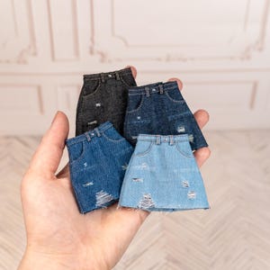 Puede incluir: Una colección de minifaldas vaqueras en varios tonos de azul y gris. Cada falda presenta detalles desgastados, incluyendo secciones rasgadas. Las faldas se sostienen en una mano, mostrando los diferentes lavados y texturas del denim.