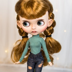 May include: A doll with long, red hair styled in braids with black hair ties. The doll is wearing a teal turtleneck sweater and blue jeans with distressed details.