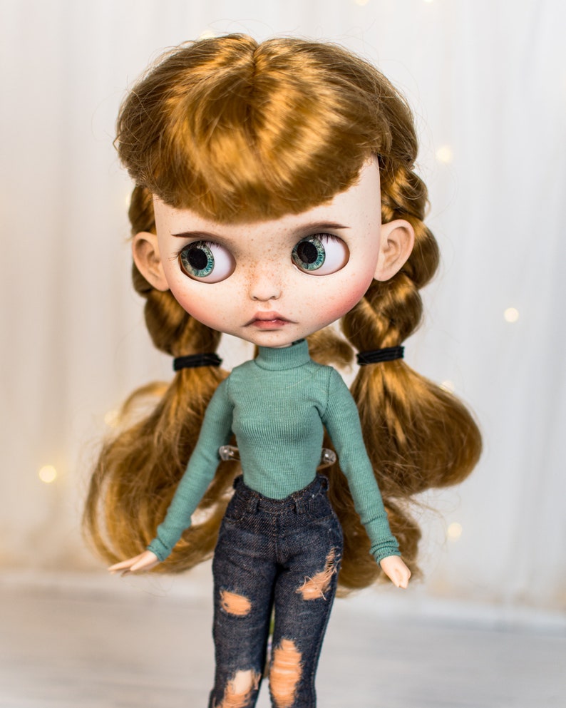 May include: A doll with long, red hair styled in braids with black hair ties. The doll is wearing a teal turtleneck sweater and blue jeans with distressed details.