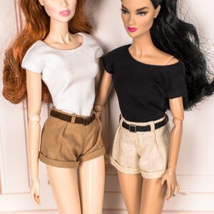 May include: Two dolls wearing different outfits. One doll is wearing a white short-sleeved shirt and brown shorts with a brown belt. The other doll is wearing a black short-sleeved shirt and beige shorts with a black belt.