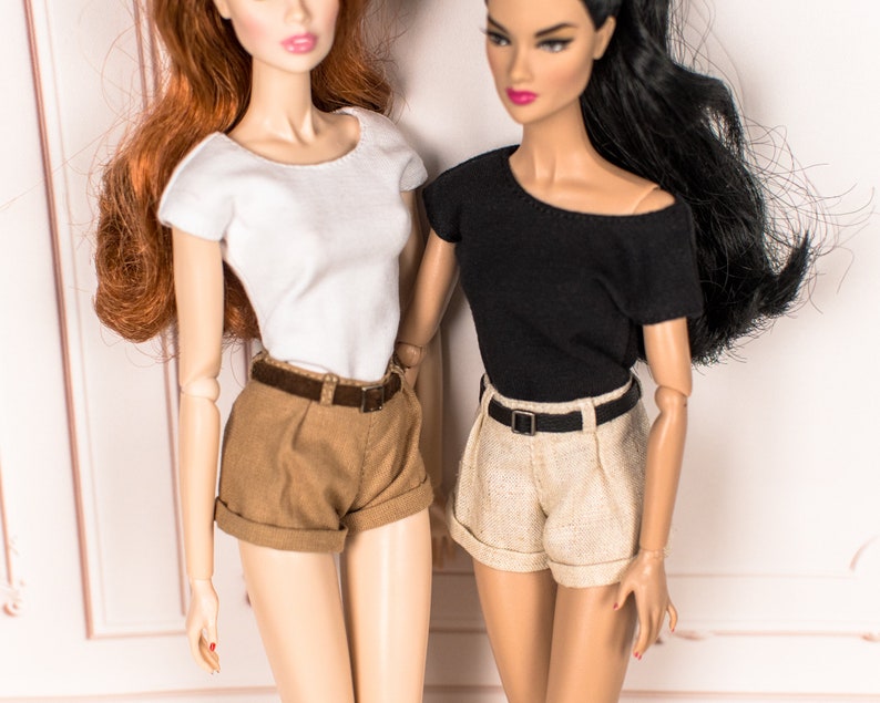 May include: Two dolls wearing different outfits. One doll is wearing a white short-sleeved shirt and brown shorts with a brown belt. The other doll is wearing a black short-sleeved shirt and beige shorts with a black belt.