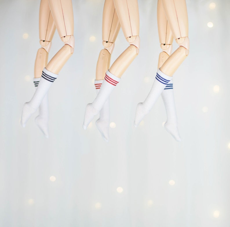May include: Three pairs of white socks with colored stripes. The socks are hanging from the legs of three dolls.