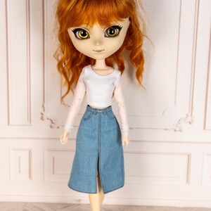 May include: A doll with auburn hair and large yellow eyes, wearing a white long-sleeved top and a light blue denim skirt with a front slit. The doll is also wearing white Mary Jane shoes.