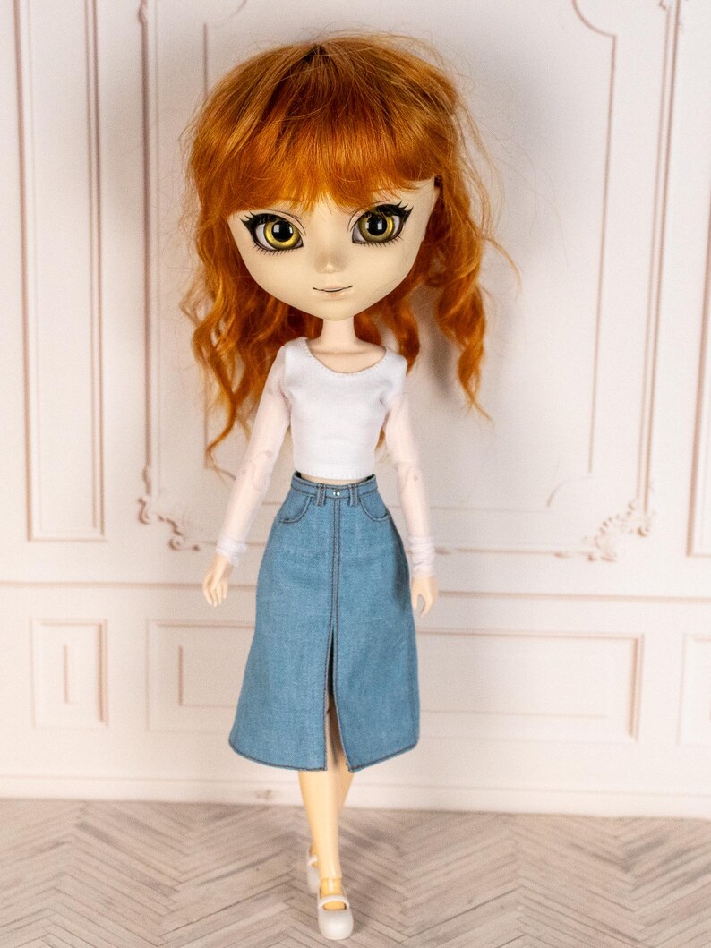 May include: A doll with auburn hair and large yellow eyes, wearing a white long-sleeved top and a light blue denim skirt with a front slit. The doll is also wearing white Mary Jane shoes.