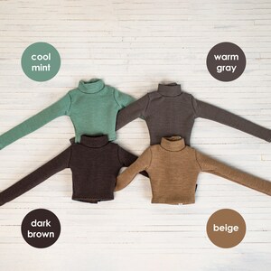 May include: Four long-sleeved turtlenecks in different colors: cool mint, warm gray, dark brown, and beige. The shirts are laid out on a white wooden surface.