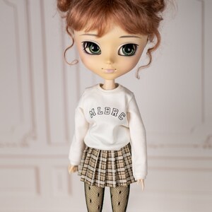 May include: A doll with brown hair in pigtails wearing a white sweatshirt with "MLBRC" printed on the front, a plaid skirt, and black tights.