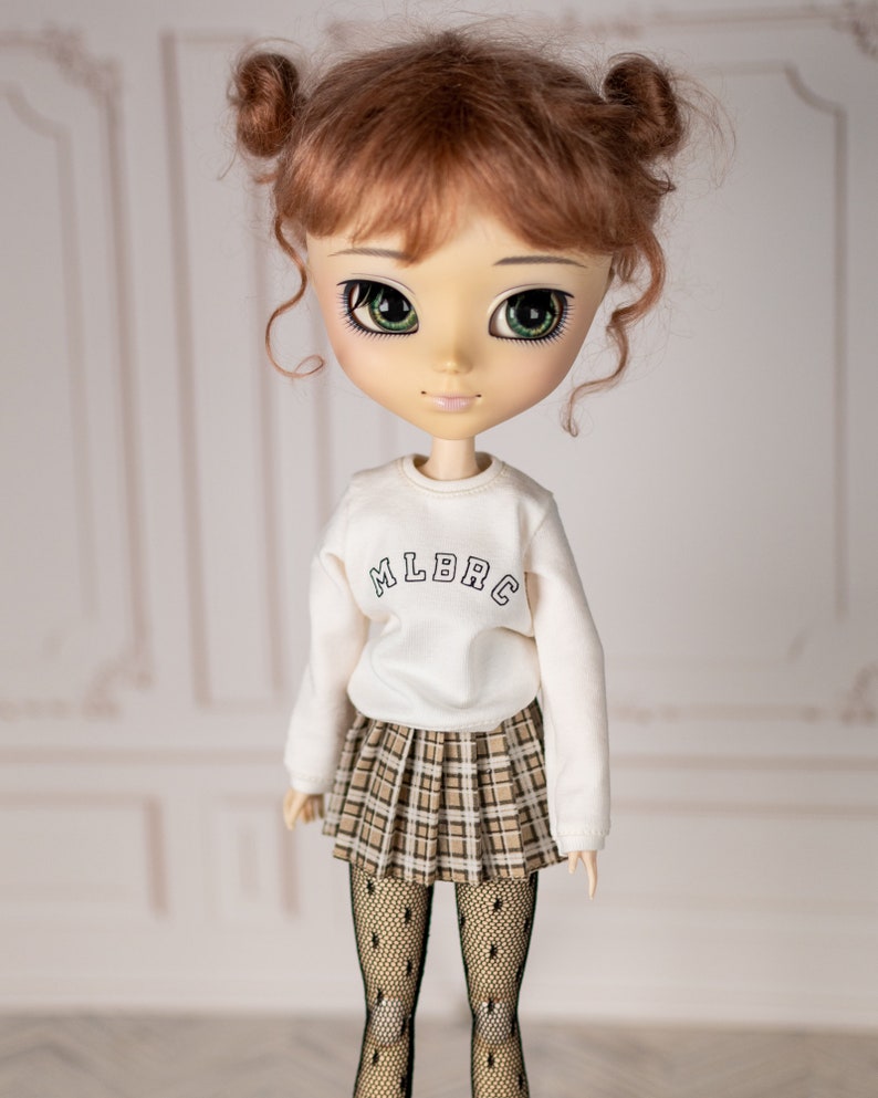 May include: A doll with brown hair in pigtails wearing a white sweatshirt with "MLBRC" printed on the front, a plaid skirt, and black tights.