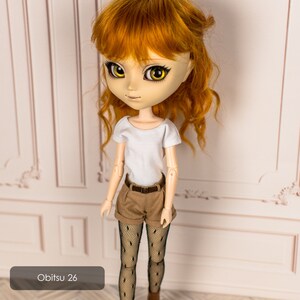 May include: A doll with red hair wearing a white t-shirt, brown shorts, and black tights. The doll is standing in front of a white wall with a decorative pattern. The text "Obitsu 26" is visible in the lower left corner of the image.