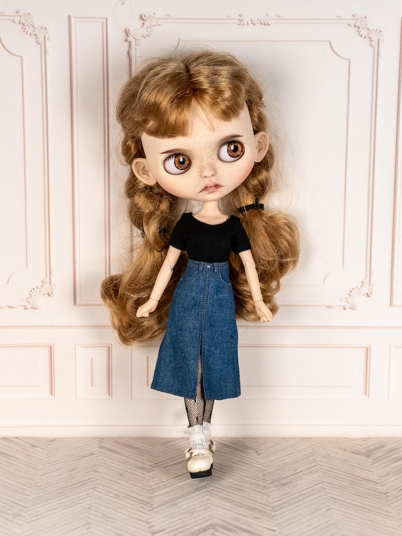 May include: A doll with long, wavy auburn hair styled in pigtails. The doll wears a black short-sleeved top and a denim skirt with a front slit. The doll's outfit is completed with white shoes and fishnet stockings.