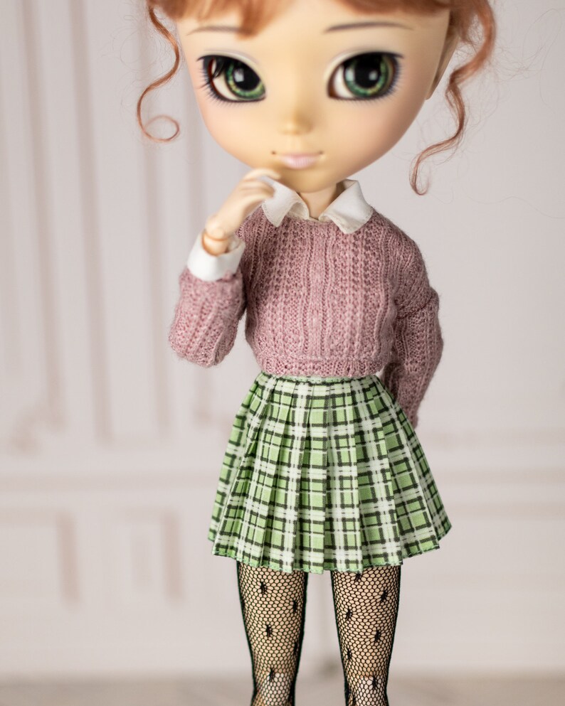 May include: A doll with large green eyes, wearing a pink sweater, a green plaid skirt, and black tights.