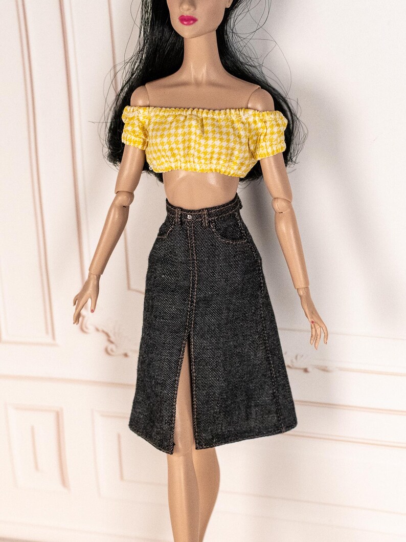 May include: A doll wearing a yellow and white gingham off-the-shoulder crop top and a dark denim skirt with a front slit. The skirt has a high waist and a visible belt loop. The outfit is designed for a doll.