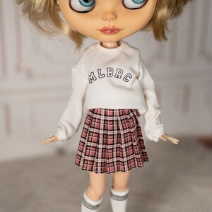 May include: A doll wearing a white long-sleeved sweatshirt with the text "MLBRC" on the front and a plaid skirt with pink, black, and white squares.