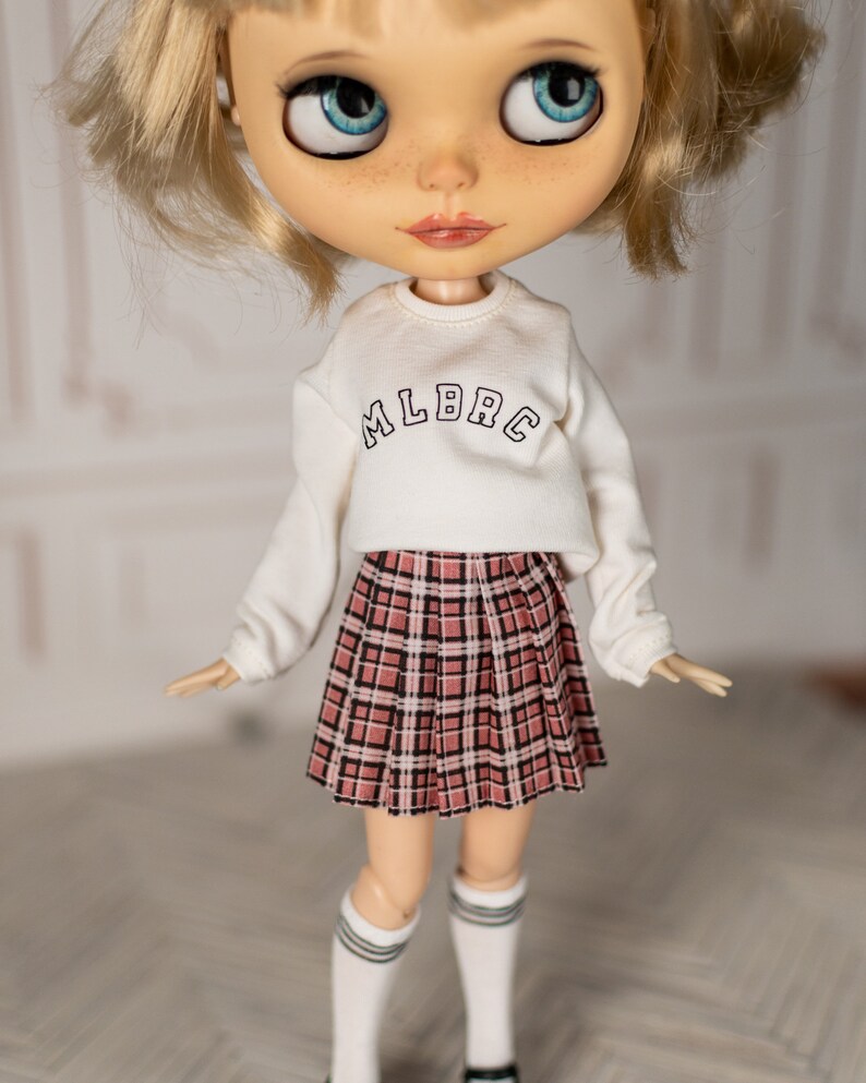 May include: A doll wearing a white long-sleeved sweatshirt with the text "MLBRC" on the front and a plaid skirt with pink, black, and white squares.