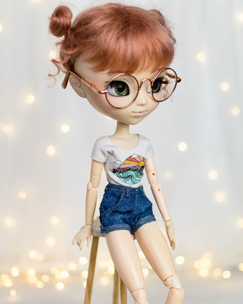 May include: A fashion doll with auburn hair styled in two buns, wearing round gold-rimmed glasses, a white t-shirt with a colorful graphic, and denim shorts. The doll is posed sitting on a small wooden stool.
