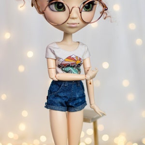 May include: A doll wearing a white t-shirt with a colorful graphic and denim shorts. The doll has red hair, round glasses, and is posed with one arm bent. The t-shirt features a sun, waves, and the text "So Bad It's Good".