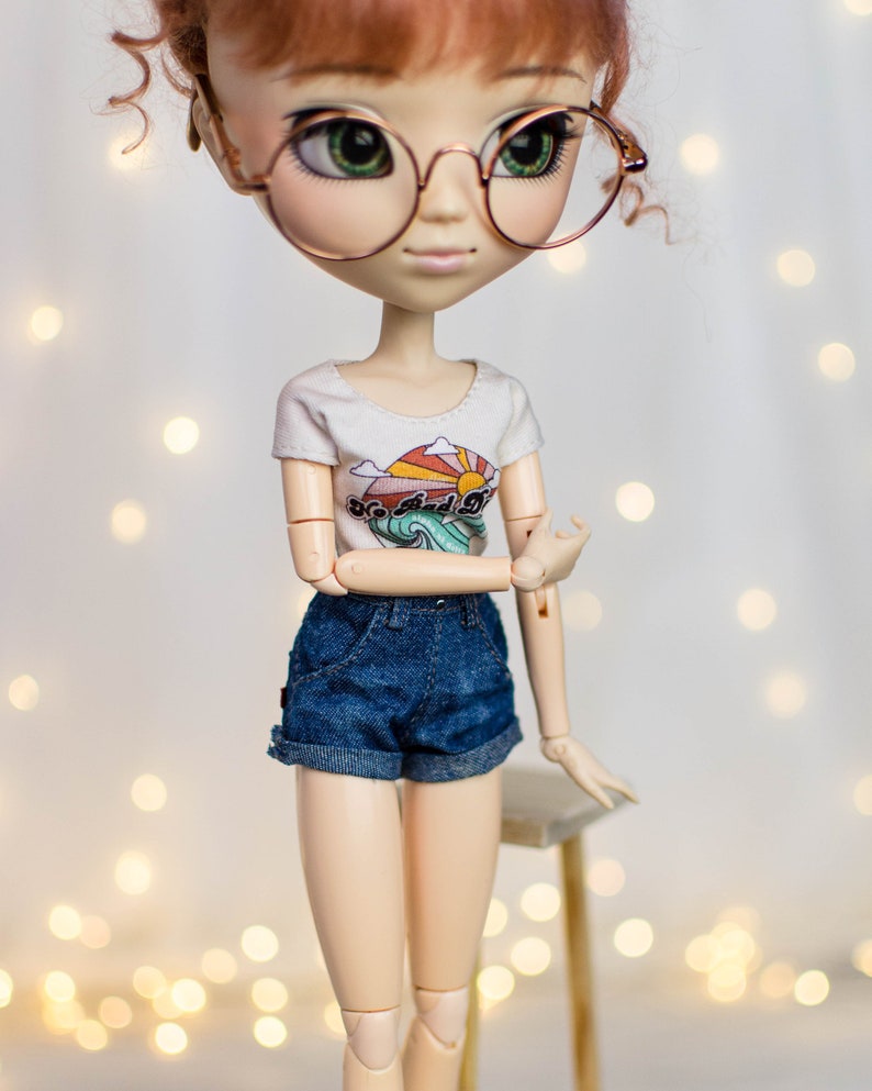 May include: A doll wearing a white t-shirt with a colorful graphic and denim shorts. The doll has red hair, round glasses, and is posed with one arm bent. The t-shirt features a sun, waves, and the text "So Bad It's Good".