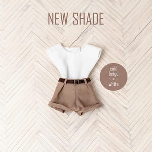 May include: A doll outfit with a white sleeveless shirt and brown shorts with a brown belt. The outfit is on a white and brown wood background. The text "NEW SHADE" is at the top of the image. The text "cold beige + white" is in a circle on the right side of the image.