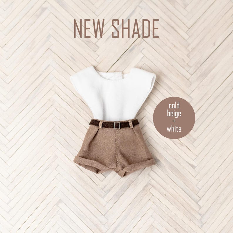 May include: A doll outfit with a white sleeveless shirt and brown shorts with a brown belt. The outfit is on a white and brown wood background. The text "NEW SHADE" is at the top of the image. The text "cold beige + white" is in a circle on the right side of the image.