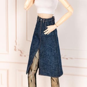 May include: A white ribbed crop top paired with a dark wash denim skirt featuring a front slit. The outfit is accessorized with black patterned tights. The doll-sized clothing is displayed against a light background.