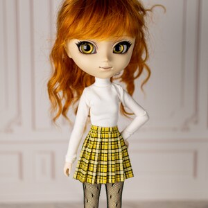 May include: A doll with long, curly red hair, wearing a white turtleneck sweater and a yellow and black plaid skirt. The doll has large, yellow eyes and a sweet expression.
