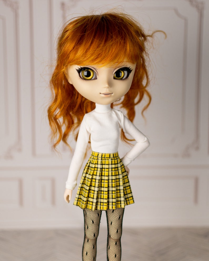 May include: A doll with long, curly red hair, wearing a white turtleneck sweater and a yellow and black plaid skirt. The doll has large, yellow eyes and a sweet expression.