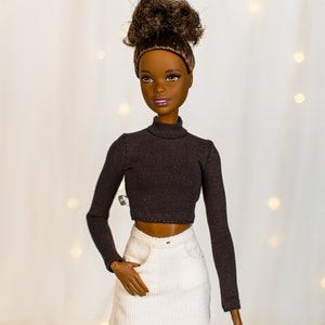May include: A doll with brown skin and brown hair styled in a bun is wearing a black turtleneck top and a white corduroy skirt.