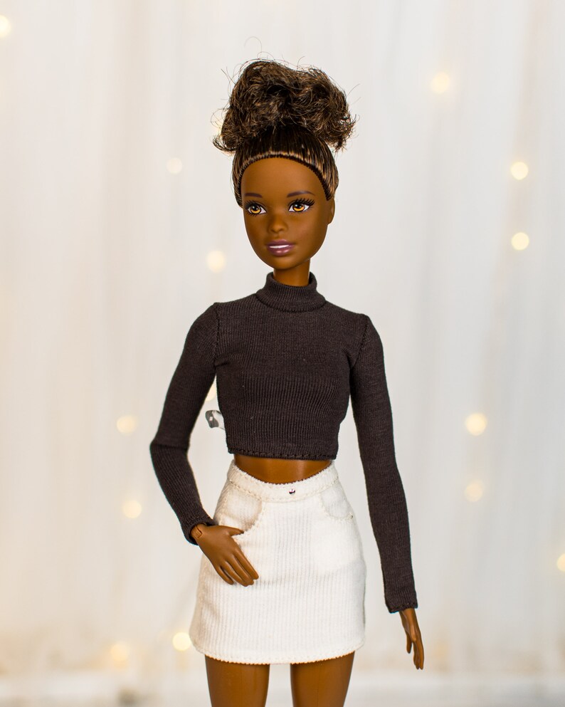 May include: A doll with brown skin and brown hair styled in a bun is wearing a black turtleneck top and a white corduroy skirt.