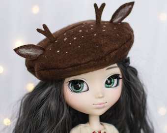 May include: A doll with long black hair wearing a brown felt beret with deer antlers. The beret has white snowflake-like designs. The doll has green eyes and is wearing a beige dress with a red bow.
