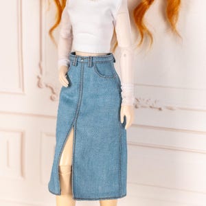 May include: A doll wearing a white long-sleeved top with sheer sleeves and a light blue denim skirt. The skirt has a front slit and is detailed with visible stitching. The doll has red hair.