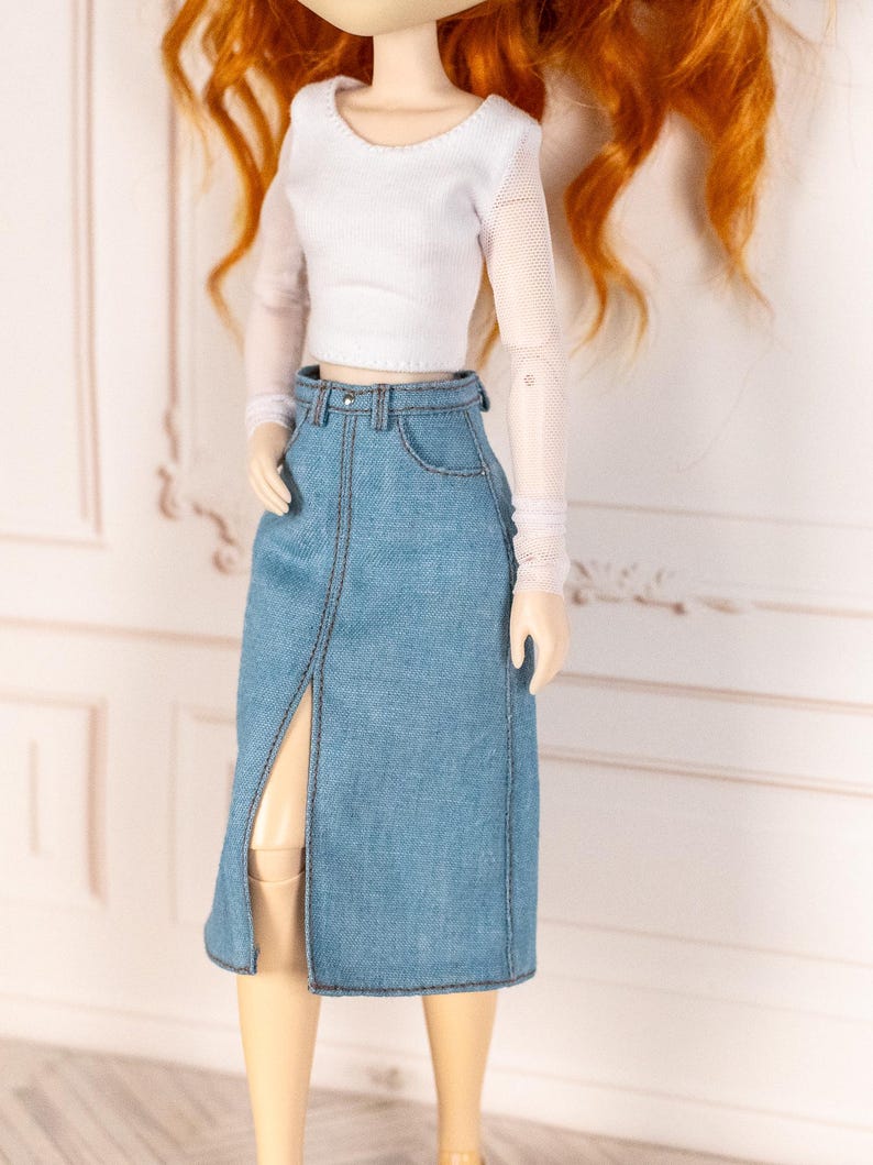 May include: A doll wearing a white long-sleeved top with sheer sleeves and a light blue denim skirt. The skirt has a front slit and is detailed with visible stitching. The doll has red hair.