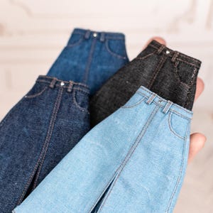 May include: A collection of miniature denim jeans in various shades of blue and black. The jeans have classic five-pocket styling with visible stitching and metal button closures. The jeans are held in a hand.