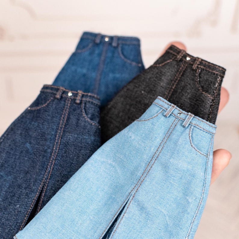 May include: A collection of miniature denim jeans in various shades of blue and black. The jeans have classic five-pocket styling with visible stitching and metal button closures. The jeans are held in a hand.